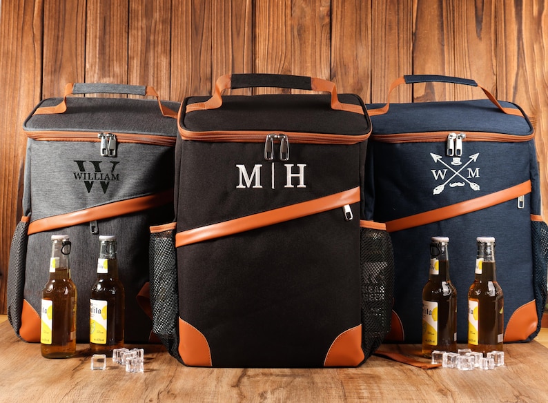 Pu&ograve; includere: Tre zaini frigo personalizzati in grigio, nero e blu navy, ciascuno con dettagli in pelle marrone. Lo zaino grigio ha il nome "WILLIAM", lo zaino nero ha le iniziali "M|H" e lo zaino blu navy ha le iniziali "WXM".