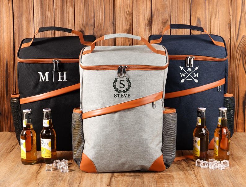 Pu&ograve; includere: Tre zaini frigo personalizzati in nero, grigio e blu navy, con dettagli in pelle marrone. Ogni borsa presenta un monogramma o un nome diverso, con bottiglie di birra e cubetti di ghiaccio davanti.