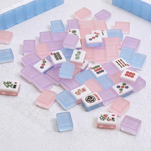 May include: A collection of pastel-colored Mahjong tiles in shades of pink, blue, and lavender, scattered on a white surface. The tiles feature various symbols and numbers, with two blue and white tile holders in the background.