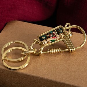 Könnte beinhalten: Ein goldfarbener Schlüsselanhänger aus Metall mit einem dekorativen Anhänger in Form eines kleinen Wagens. Der Wagen ist mit bunten Perlen gefüllt und hat eine Feder daran befestigt. Der Schlüsselanhänger hat einen Doppelring zum Anbringen von Schlüsseln.