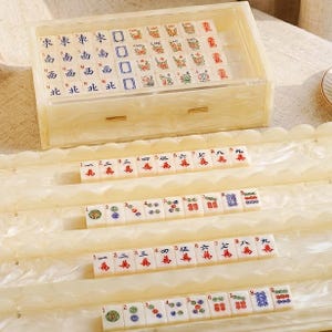 May include: A mahjong set featuring off-white tiles with colorful Chinese characters and symbols, housed in a rectangular, pearlescent cream-colored acrylic case with a drawer. Four matching scalloped racks hold rows of tiles in front of the case.
