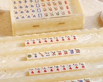 May include: A mahjong set featuring off-white tiles with colourful Chinese characters and symbols, housed in a rectangular, pearlescent cream-coloured acrylic case with a drawer. Four matching scalloped racks hold rows of tiles in front of the case.