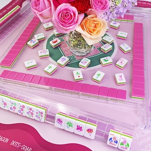 K&ouml;nnte beinhalten: Ein rosa-wei&szlig;es Mahjong-Set mit einem transparenten Acrylgeh&auml;use. Die Spielsteine sind wei&szlig; mit farbenfrohen Symbolen und gr&uuml;nen und rosa Akzenten. Eine Vase mit rosa und orangefarbenen Rosen steht in der Mitte des Spielbretts.