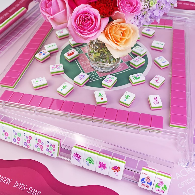 K&ouml;nnte beinhalten: Ein rosa-wei&szlig;es Mahjong-Set mit einem transparenten Acrylgeh&auml;use. Die Spielsteine sind wei&szlig; mit farbenfrohen Symbolen und gr&uuml;nen und rosa Akzenten. Eine Vase mit rosa und orangefarbenen Rosen steht in der Mitte des Spielbretts.