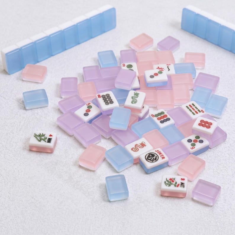 May include: A collection of pastel-colored Mahjong tiles in shades of pink, blue, and lavender, scattered on a white surface. The tiles feature various symbols and numbers, with two blue and white tile holders in the background.