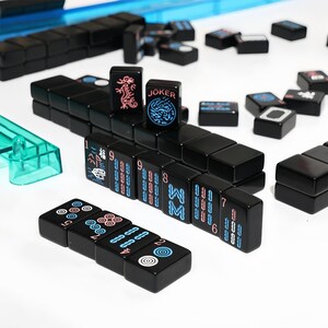 May include: A black Mahjong set with tiles displaying various symbols and numbers in white, blue, and pink. The set includes tiles marked "JOKER" and a dragon symbol. The game pieces are arranged on a white surface.