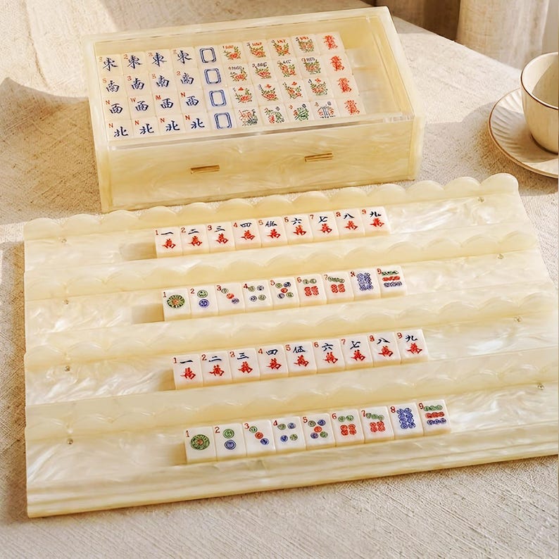 May include: A cream-colored marbled acrylic Mahjong set featuring a storage box with a drawer and four matching tile racks. The set includes white tiles with red, blue, and green engraved Chinese characters, numbers, and symbols arranged in rows.
