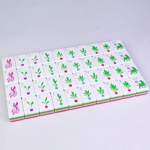 K&ouml;nnte beinhalten: Ein Satz wei&szlig;er Mahjong-Steine mit verschiedenen Symbolen und Zahlen. Die Steine zeigen Bilder von Pflanzen in T&ouml;pfen, Ph&ouml;nixen und dem Wort "BAM". Die Kanten sind mit Rosa und Gr&uuml;n verziert.