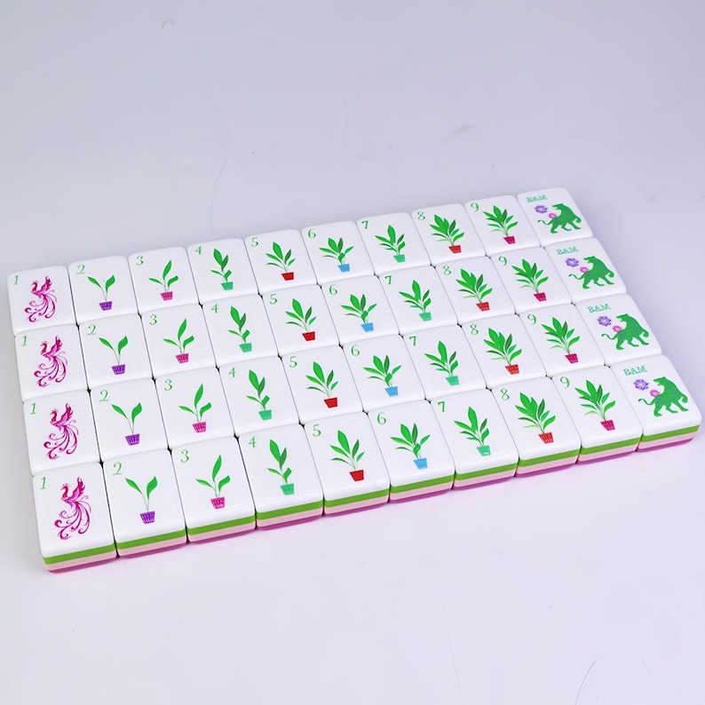 K&ouml;nnte beinhalten: Ein Satz wei&szlig;er Mahjong-Steine mit verschiedenen Symbolen und Zahlen. Die Steine zeigen Bilder von Pflanzen in T&ouml;pfen, Ph&ouml;nixen und dem Wort "BAM". Die Kanten sind mit Rosa und Gr&uuml;n verziert.