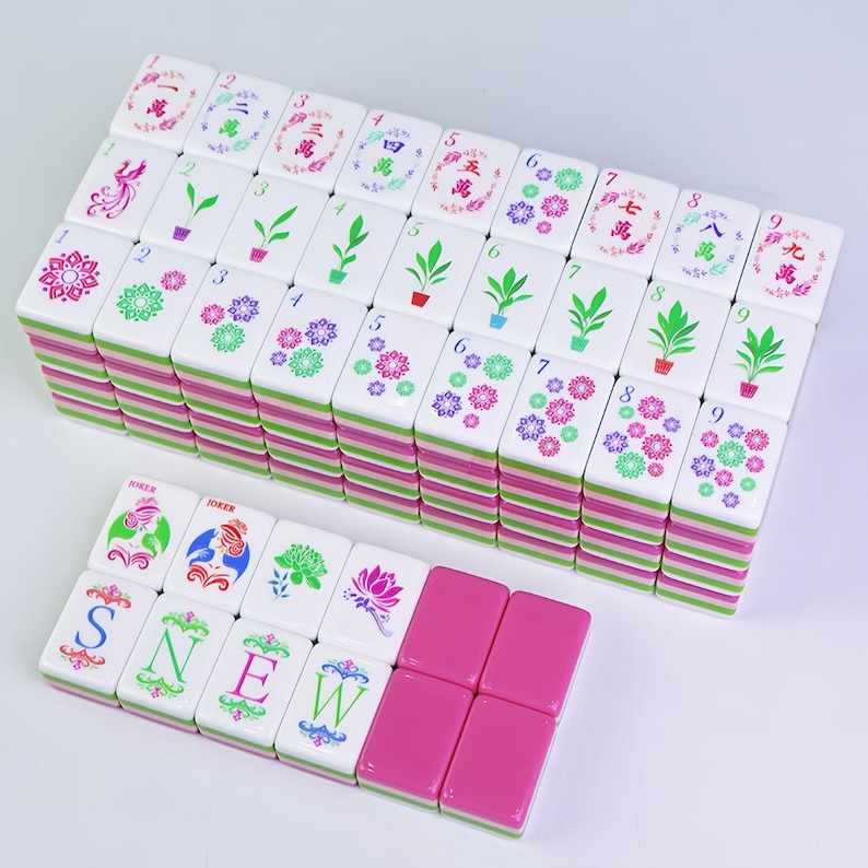 K&ouml;nnte beinhalten: Ein Satz wei&szlig;er Mahjong-Steine mit rosa und gr&uuml;nen R&auml;ndern. Die Steine zeigen verschiedene Symbole, Zahlen und florale Designs. Einige Steine schreiben "SNEW" und enthalten "JOKER"-Steine. Das Set ist auf einer wei&szlig;en Oberfl&auml;che angeordnet.