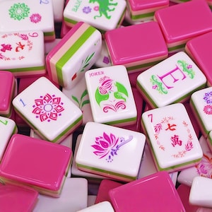 Könnte beinhalten: Eine Sammlung farbenfroher Mahjong-Steine. Die Steine weisen verschiedene Designs auf, darunter Blumenmuster, Zahlen und chinesische Schriftzeichen. Die Steine sind weiß mit rosa, grünen und anderen Farbakzenten. Einige Steine tragen das Wort "Joker".