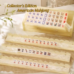 May include: A Collector's Edition American Mahjong set featuring cream-colored, marbled resin tiles and matching racks. The set includes a storage box filled with tiles, and three racks displaying various numbered and patterned game tiles.