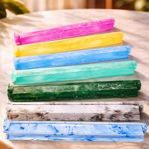 May include: A collection of seven rectangular plastic bag sealing clips arranged in a stack. The clips feature various colors including bright pink, yellow, light blue, teal, dark green, a white and black marble pattern, and a light blue floral pattern.