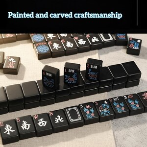 May include: A close-up of a black Mahjong set with painted and carved details. The tiles feature Chinese characters, floral designs, and numbers. The text "Painted and carved craftsmanship" is visible.