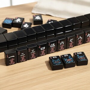 May include: Black Mahjong tiles with red and white Chinese characters and numbers. Some tiles have floral and graphic designs. The tiles are arranged on a light wood surface.