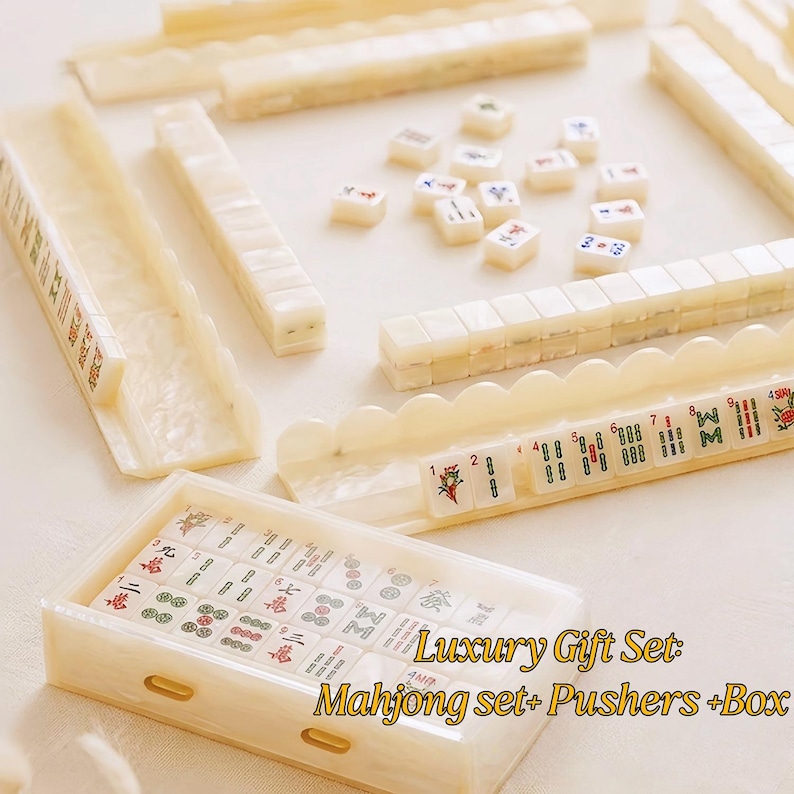 May include: A Mahjong set featuring off-white tiles with colorful symbols, characters, and numbers. The set includes matching tile racks, pushers, and a storage box. The text Luxury Gift Set: Mahjong set+ Pushers +Box is visible.