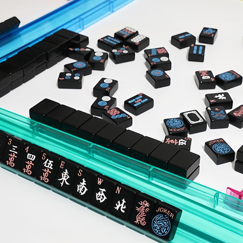 May include: A Mahjong set with black tiles and a blue plastic tray. The tiles feature various symbols and characters in white, red, and blue. Some tiles have the word "JOKER" on them.
