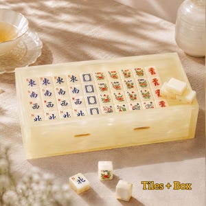 May include: A cream-colored, marbled resin box containing a set of Mahjong tiles. The tiles feature blue directional characters, green floral motifs, and red dragon symbols. Three loose tiles are placed in front of the box, and the text Tiles + Box is visible in the bottom right corner.
