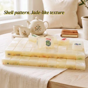 May include: A set of rectangular mahjong tiles with a cream-colored, jade-like texture and shell pattern. The tiles feature various engraved symbols and characters in red, green, and blue, including one tile clearly marked with the text JOKER.