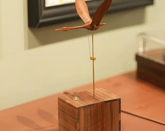 May include: A wooden kinetic sculpture of a bird in flight. The bird is made of light brown wood and is suspended above a wooden box base. A thin metal rod and wire support the bird, creating a sense of movement and balance. The base has a small silver knob.
