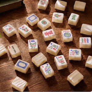 May include: A collection of off-white mahjong tiles scattered on a wooden surface. The tiles feature various colorful engraved designs, including floral patterns, numbers, and text such as 1 SPR, 3 AUT, 4 WIN, 2 SUM, and JOKER, with some tiles left blank.