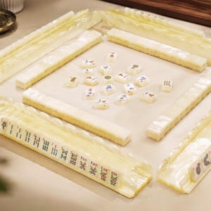 May include: A mahjong set featuring four translucent, pearlescent off-white tile racks arranged in a square. The racks hold rectangular tiles with various green, red, and blue character and symbol prints. Additional loose tiles are scattered in the center.