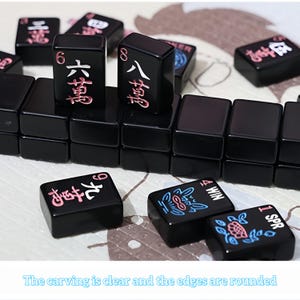 May include: Black Mahjong tiles with red and white Chinese characters and symbols. The tiles are arranged on a light-colored surface. The text "The carving is clear and the edges are rounded" is at the bottom.