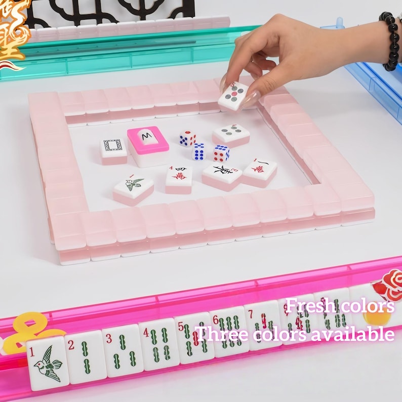 May include: A pink Mahjong set with white tiles and dice. The set includes a pink frame and a tray with tiles featuring various symbols and characters. The image also shows a tray with numbered tiles and the text "Fresh colors, three colors available."