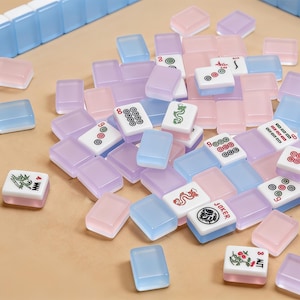 May include: A collection of pastel-colored Mahjong tiles in shades of pink, lavender, and blue, scattered on a light brown surface. Some tiles feature symbols and characters, while others are solid colors. A few rows of blue tiles are arranged at the top.
