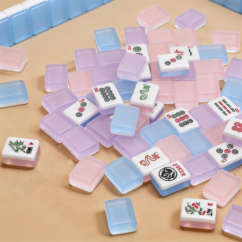 May include: A collection of pastel-colored Mahjong tiles in shades of pink, lavender, and blue, scattered on a light brown surface. Some tiles feature symbols and characters, while others are solid colors. A few rows of blue tiles are arranged at the top.