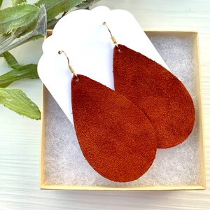 May include: A pair of teardrop-shaped earrings in a rich, reddish-brown suede texture. The earrings are displayed in a small, beige gift box with a white backing and gold-colored ear wires. The earrings are approximately 3 inches long.