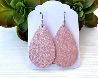 May include: A pair of teardrop-shaped earrings in a light pink color. The earrings have a textured surface and are attached to silver-colored ear wires. The earrings are displayed on a white card.