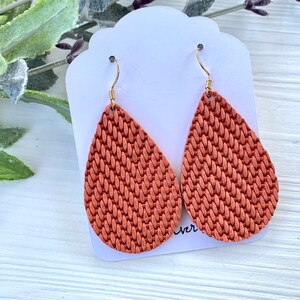 May include: A pair of teardrop-shaped earrings with a woven texture in a warm, terracotta color. The earrings hang from gold-colored hooks and are displayed on a white card with a scalloped edge. The background includes greenery.