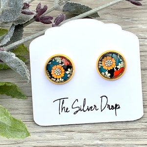 May include: Gold-toned stud earrings with a floral design. The earrings feature a round, colorful floral pattern set within a gold frame. The text "The Silver Drop" is printed on the white card.