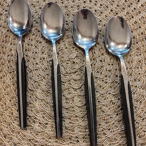 May include: Four stainless steel spoons with black handles are arranged on a woven, tan surface. The spoons have a classic design, with a polished, oval bowl and a sleek, black handle. The spoons are likely for eating.