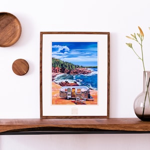 May include: A framed painting of a silver Airstream trailer parked on a beach with a view of the ocean and a rocky coastline. The sky is blue with white clouds. The painting is in a brown frame and is hanging on a white wall.