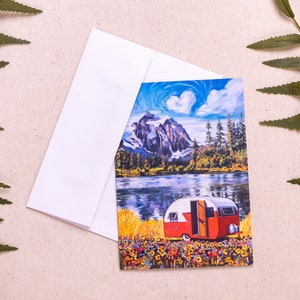 Puede incluir: Una tarjeta de felicitación con una pintura de una caravana vintage roja y blanca estacionada en un campo de flores silvestres, con un lago y montañas en el fondo. El cielo es azul con una nube en forma de corazón.