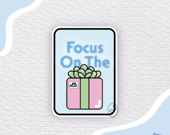 Puede incluir: Una pegatina con fondo azul claro y borde blanco. La pegatina presenta una caja de regalo rosa con una cinta verde y una pequeña planta verde en la parte superior. El texto "Focus On The" está escrito en azul sobre la caja de regalo.