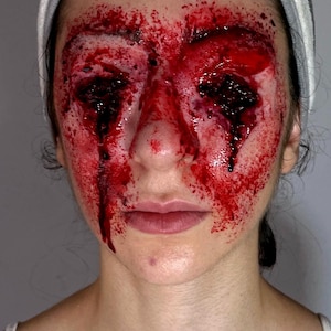 May include: A close-up of a face with realistic special effects makeup simulating severe injuries. The makeup features extensive red and dark red coloring around the eyes, forehead, and cheeks, creating a bloody and gruesome appearance. A white headband is visible.
