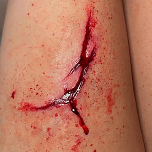 May include: A realistic special effects makeup application depicting a Y-shaped laceration on skin. The wound features deep red, glossy simulated blood pooling within the centre of the cut, surrounded by scattered red droplets and splatter marks.