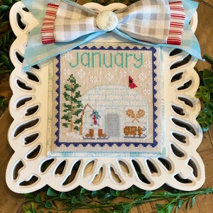 May include: A white framed cross-stitch with a blue and white checkered ribbon bow. The cross-stitch depicts a snowy scene with an igloo, a cardinal, a pine tree, and the word "January".
