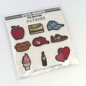 May include: A set of Steve Madden peel and stick patches in a clear plastic package. The patches include a red apple, a credit card, a blue cap, a hot dog, a red hat, a sneaker, an ice cream cone, a lipstick, and two hearts. Text reads: STEVE MADDEN PEEL AND STICK PATCHES CUSTOMIZE YOUR BAG.