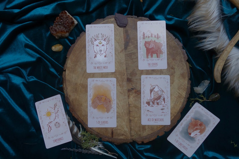 Op de afbeelding: Een spreiding van tarotkaarten op een houten oppervlak met een turquoise fluwelen achtergrond. De kaarten omvatten "Temperance", "The Lovers", "The White Wolf", "5 of Leaders", "Ace of Watchers" en "5 of Caretakers".