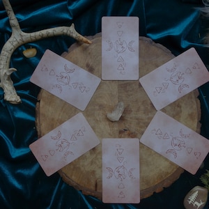 Op de afbeelding: Een set van zes tarotkaarten met een maan- en driehoeksontwerp op een houten oppervlak. De kaarten zijn in een cirkel rond een kristal gerangschikt.