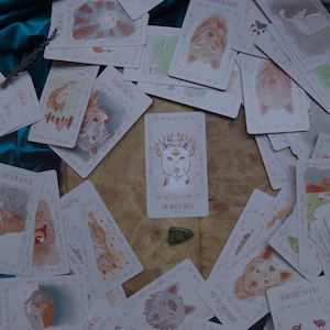 Op de afbeelding: Een spreiding van kaarten met illustraties van wolven, met de kaart "De Witte Wolf" in het midden. De kaarten liggen op een houten oppervlak met een groene steen in het midden.