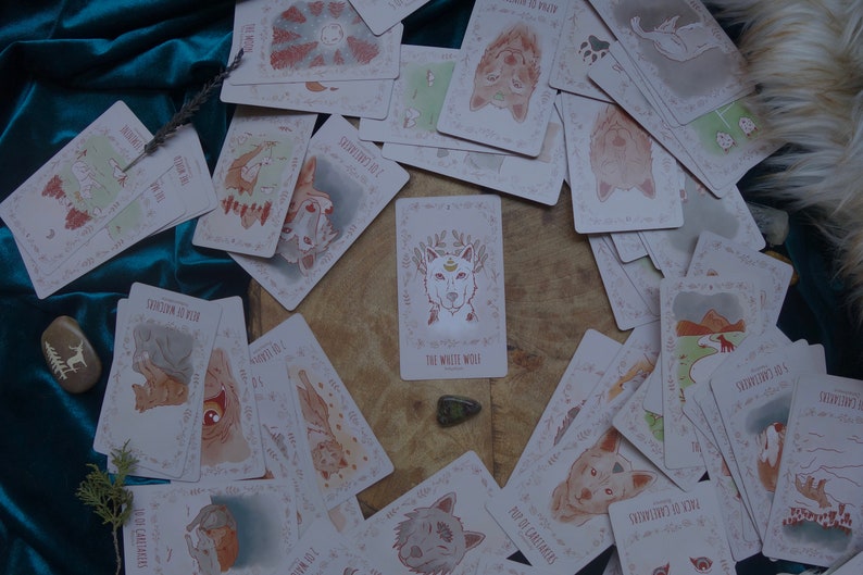 Op de afbeelding: Een spreiding van kaarten met illustraties van wolven, met de kaart "De Witte Wolf" in het midden. De kaarten liggen op een houten oppervlak met een groene steen in het midden.
