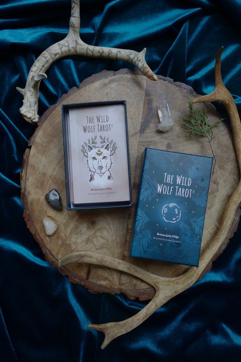 Op de afbeelding: Een blauwe doos met de tekst "The Wild Wolf Tarot" en de naam "Briana Jolie Hillje" erop. De doos staat op een houten oppervlak met geweien en kristallen.