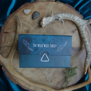 Op de afbeelding: Een zwarte doos met de tekst "The Wild Wolf Tarot" op de bovenkant. De doos staat op een houten oppervlak met geweien en stenen.