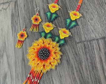 May include: A beaded jewellery set consisting of a necklace and two earrings. The necklace features a large sunflower pendant with yellow, orange, and black beads, accented by smaller floral motifs and a long fringe of orange and yellow beads.