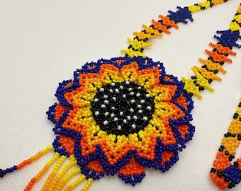 May include: A beaded necklace featuring a large, circular flower-shaped pendant made of small, vibrant yellow, orange, blue, and black beads. The central part of the flower is black with white accents, and the chain consists of matching beaded strands.